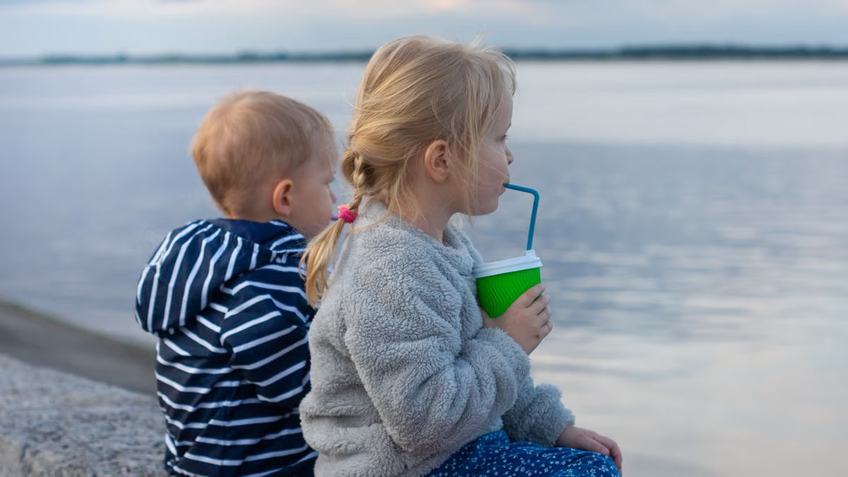Two children sit by a lake, one drinking from a cup, enjoying a peaceful day outdoors.